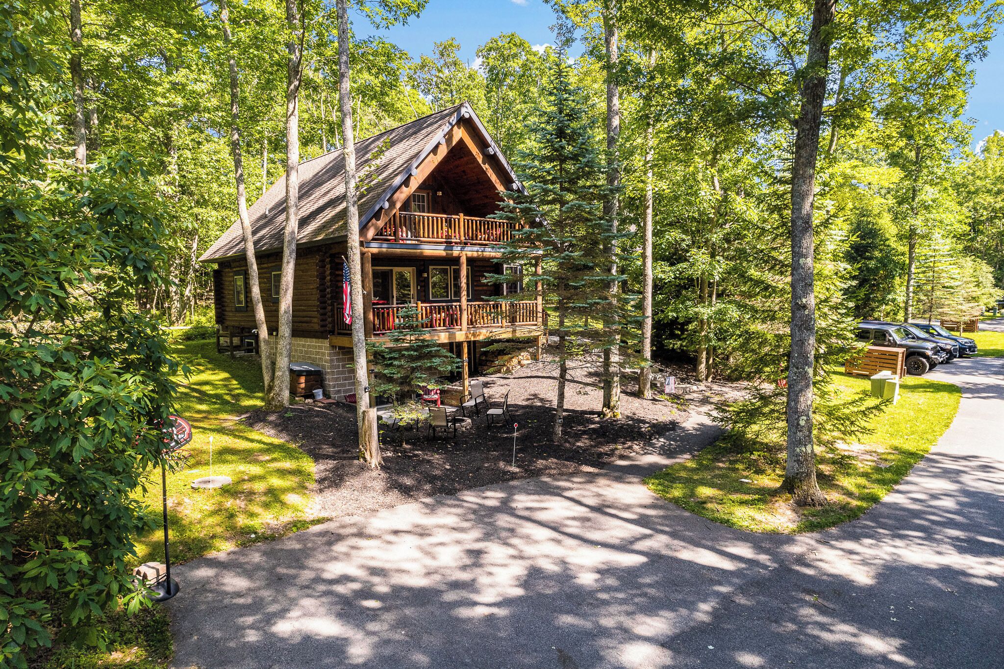 A log cabin with a balcony, surrounded by trees and a paved driveway.