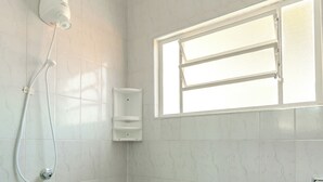 Un baño con baldosas de mármol blanco, un cabezal de ducha de mano, un dispensador de jabón en la pared y una ventana con persianas.