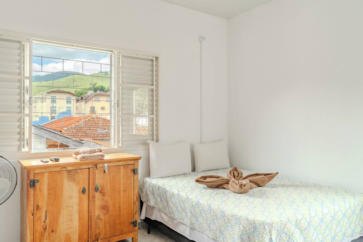 A bedroom with a bed, a wooden dresser, a window with a view of buildings and hills, and a ceiling fan.