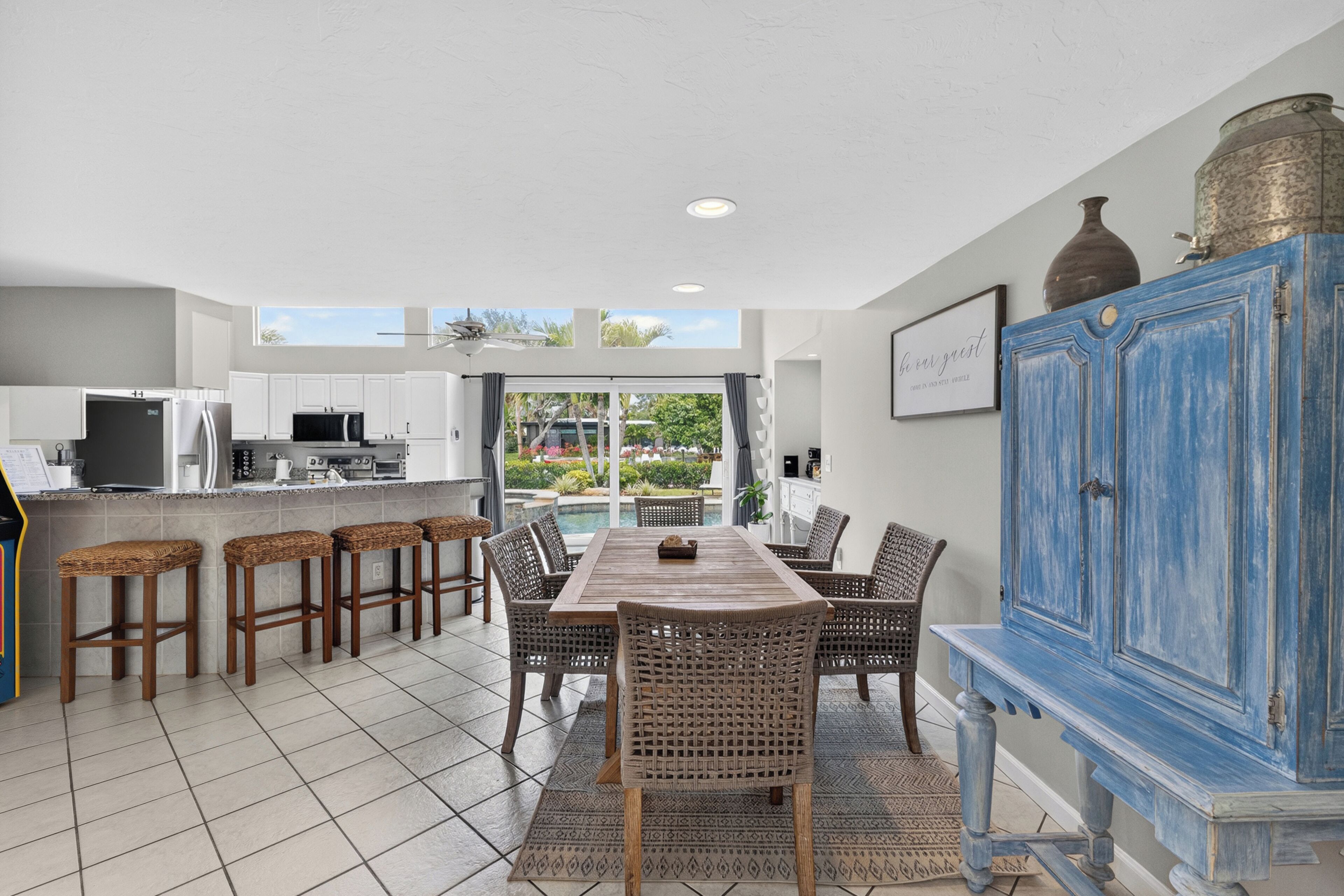 A modern kitchen with white cabinets, a dining area with a wooden table and wicker chairs, and a blue cabinet with decorative items.