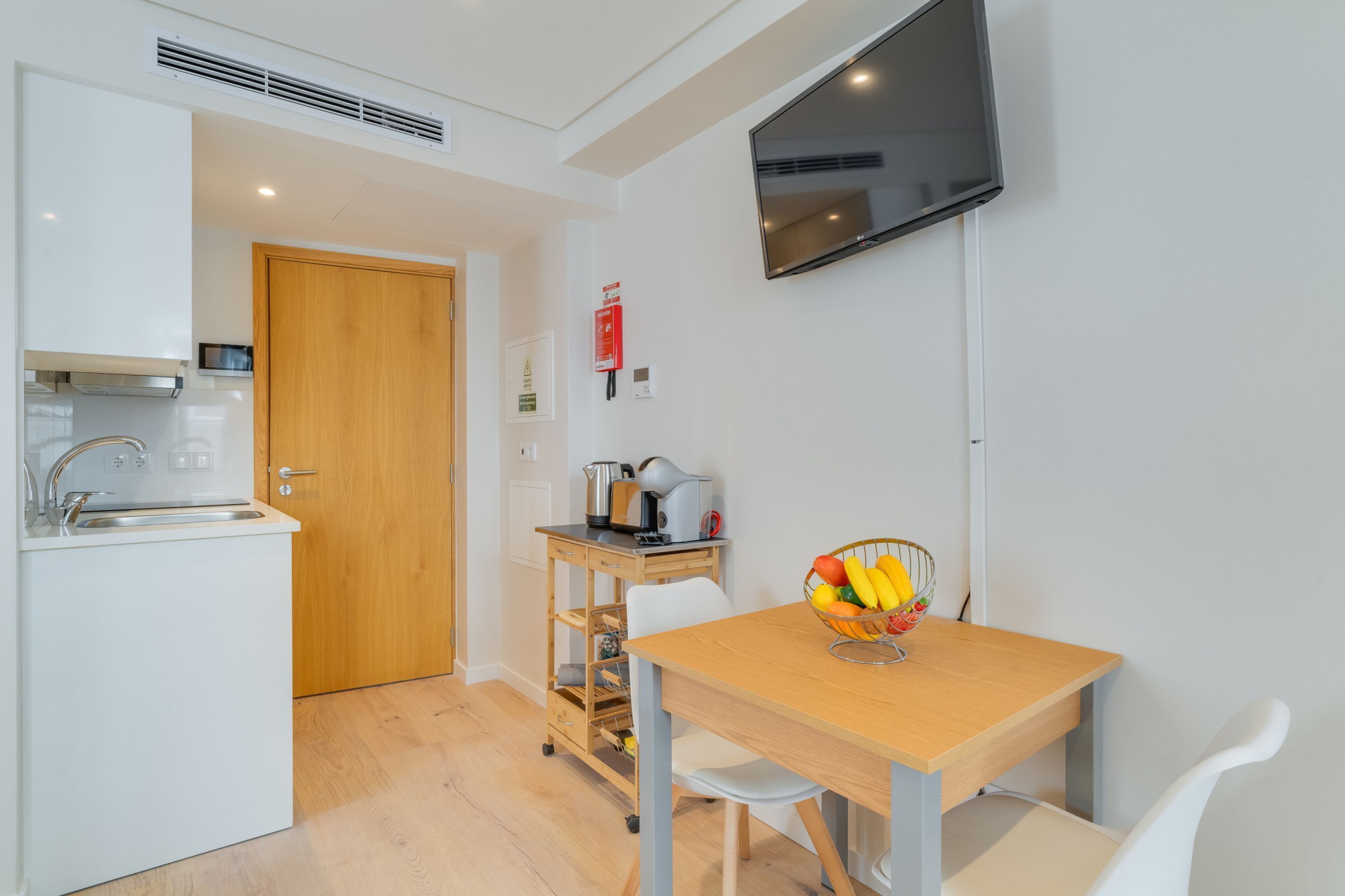 A compact kitchen with a wooden door, a white sink, a small coffee station, and a fruit bowl on a table.