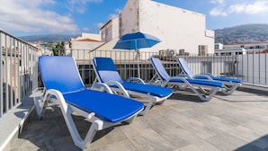 Terraza en la azotea con tumbonas y una sombrilla azul, con vistas a un paisaje urbano y montañas.