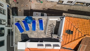Vista aérea de una azotea con sillas de descanso azules, un edificio blanco con tejado de tejas y una escalera que lleva a un nivel inferior.