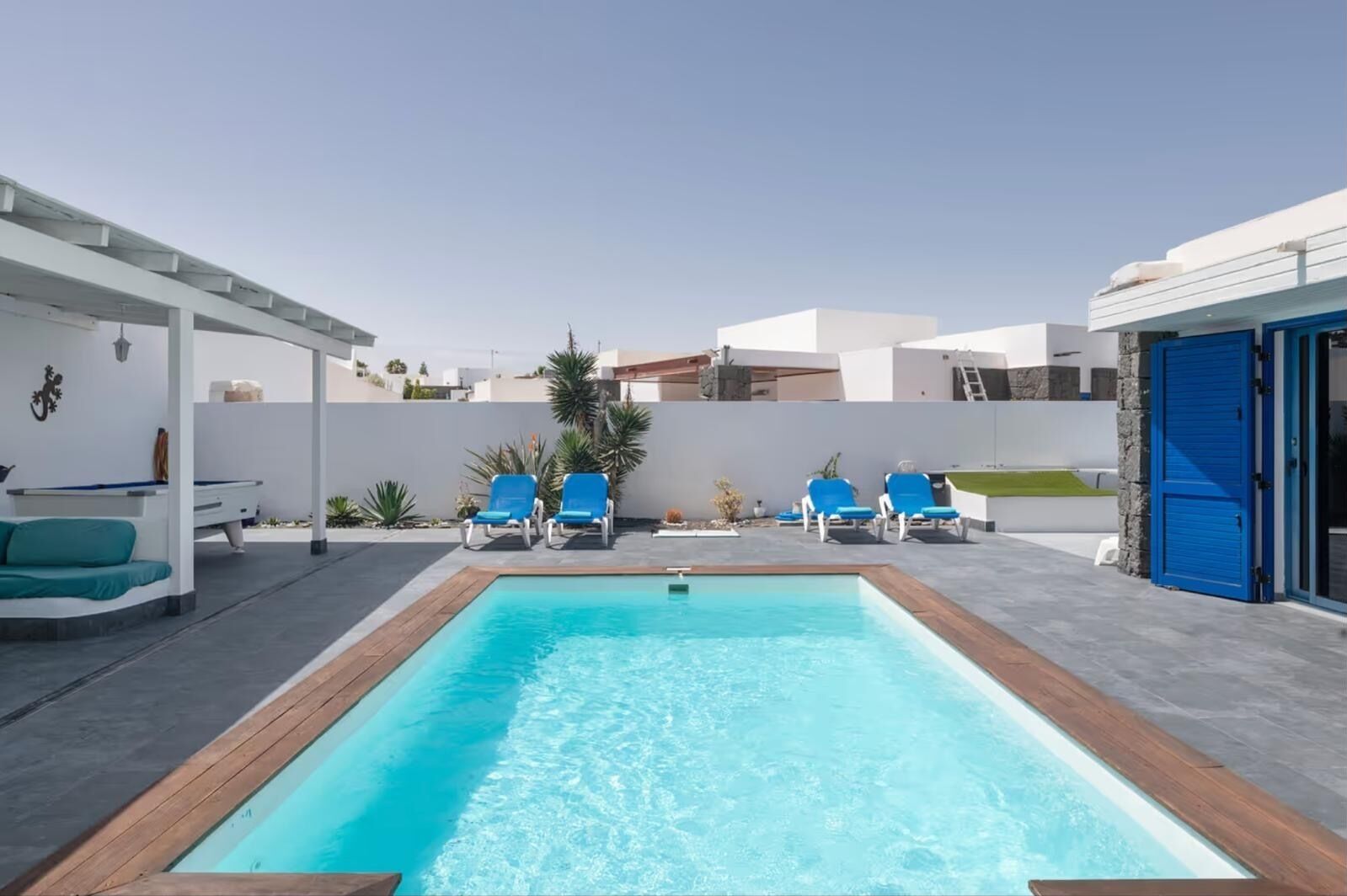 En rektangulær swimmingpool med en træterrasse langs kanten, omgivet af en hvid mur og en stenvæg. Fire blå liggestole står i række langs muren, og der er et lille siddeområde med en turkis sofa og et poolbord.