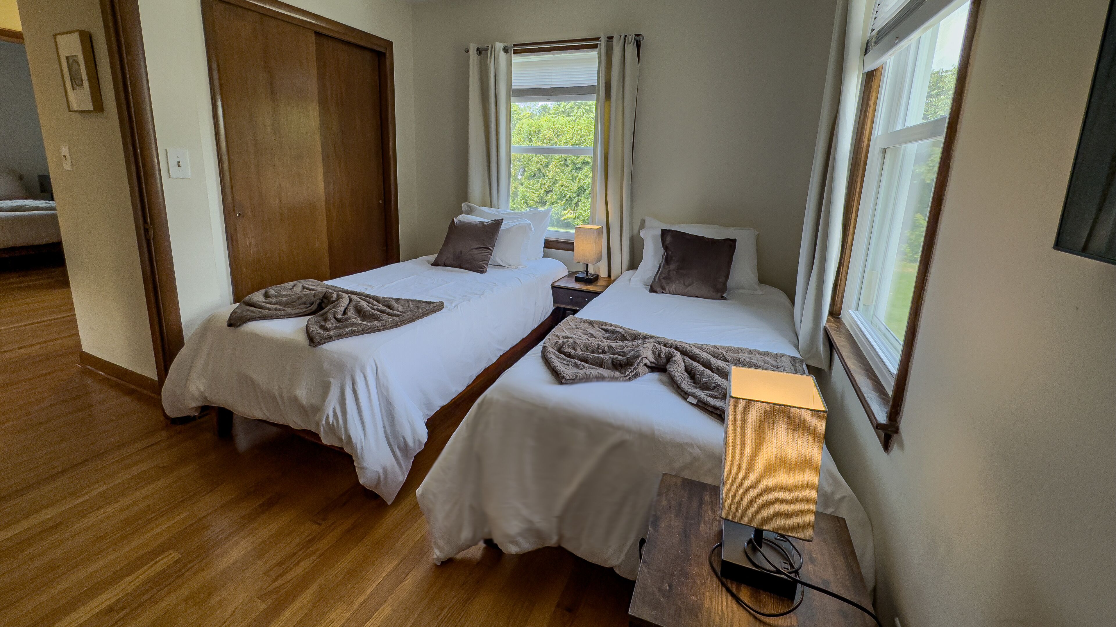 A room with two single beds, a wooden floor, a window with curtains, and a bedside lamp.