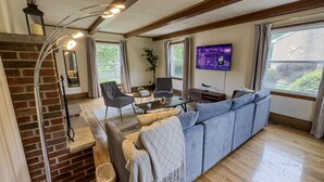 A spacious living room with a brick fireplace, a large grey sectional sofa, a coffee table, and a flat-screen TV mounted on the wall. - Elegant 4BR Princeton Escape • Game Rm • Big Yard (Princeton)