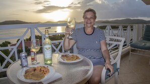 Una persona sentada a una mesa con un plato de comida, una copa de vino y una botella de Cuvée. La mesa está en un balcón al aire libre con vistas a un cuerpo de agua y montañas al fondo.