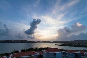 Vista costeira com um corpo de água, colinas distantes e edifícios com telhados de barro avermelhado.