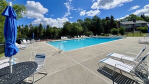 Ein Swimmingpool mit Liegestühlen und Sonnenschirmen, umgeben von Bäumen und blauem Himmel.