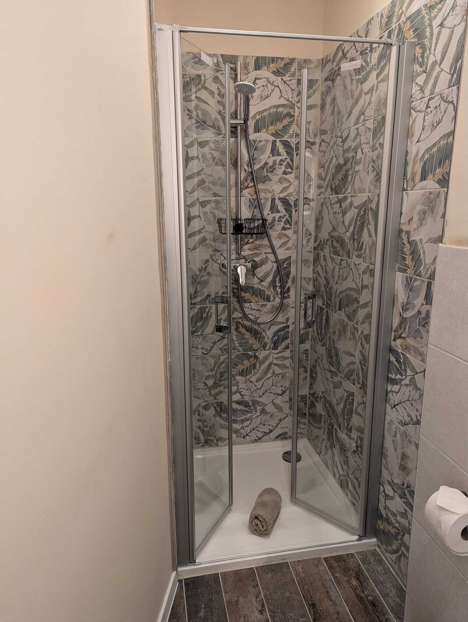Eine durch eine Glaswand abgetrennte Duschkabine mit einer Fliesenwand im Blattmuster, ein Handtuch auf dem Boden und ein Toilettenpapierhalter auf der rechten Seite.