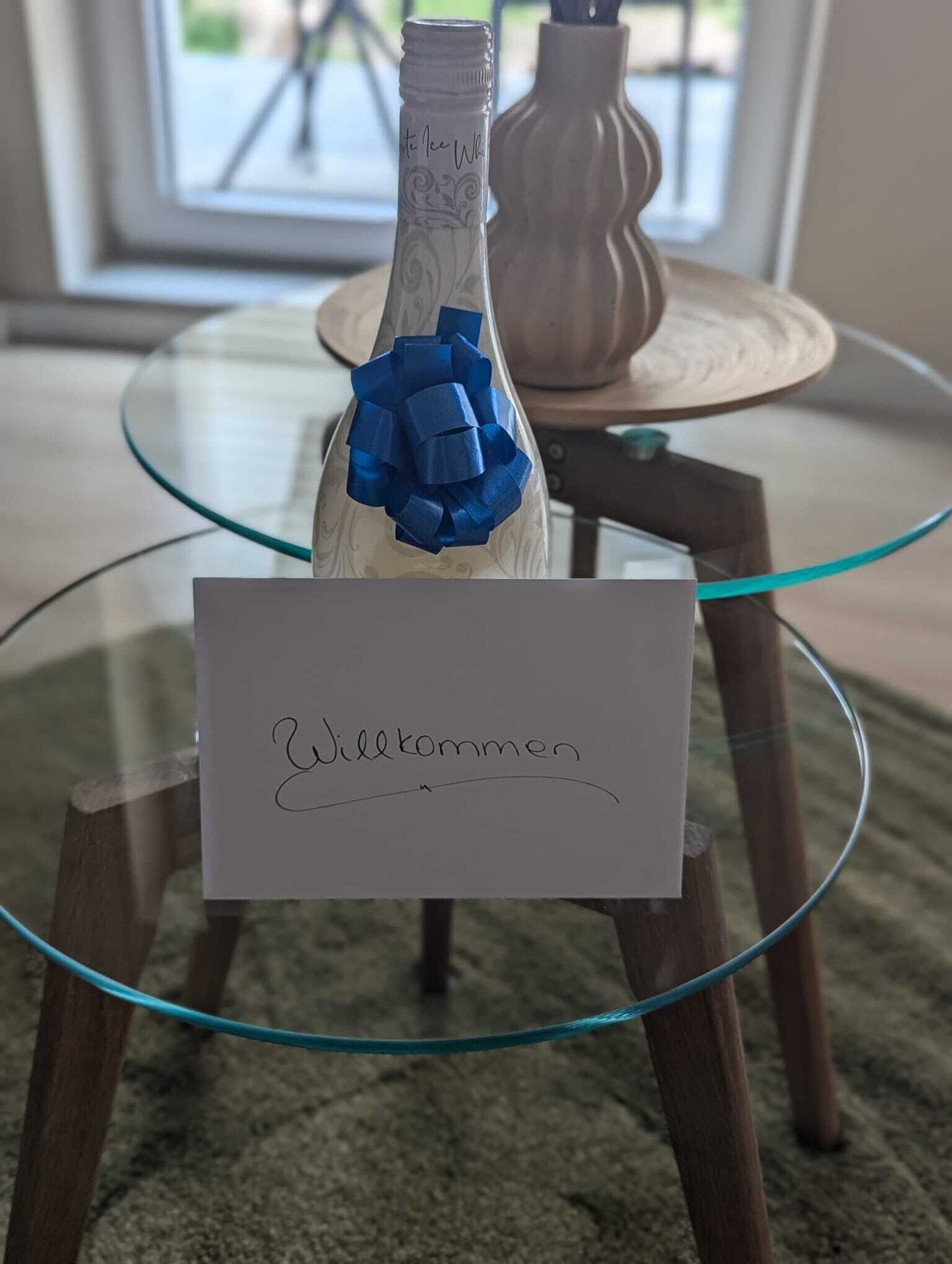 Une bouteille ornée d’un ruban bleu, posée sur une table en verre, à côté d’une pancarte indiquant « Bienvenue ».