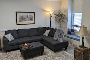 A living room with a sectional sofa, a small ottoman, a floor lamp, a potted plant, and a window with blinds.