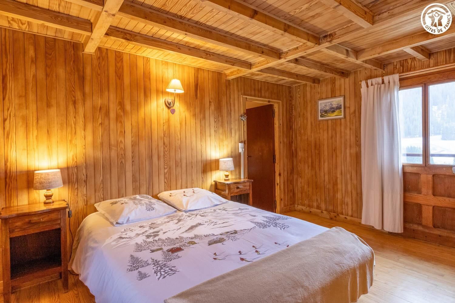 Une pièce aux murs lambrissés, comprenant un lit, deux tables de chevet avec des lampes, une fenêtre avec des rideaux et une porte.