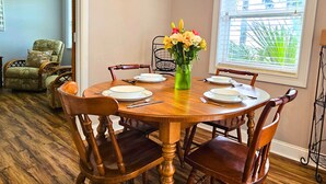 A dining room with a wooden table set for four, surrounded by wooden chairs. A vase with flowers is on the table, and a window with blinds is visible in the background.