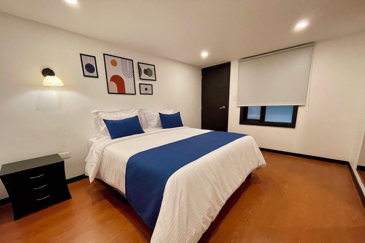 A bedroom with a bed, a nightstand, a wall-mounted lamp, and artwork.