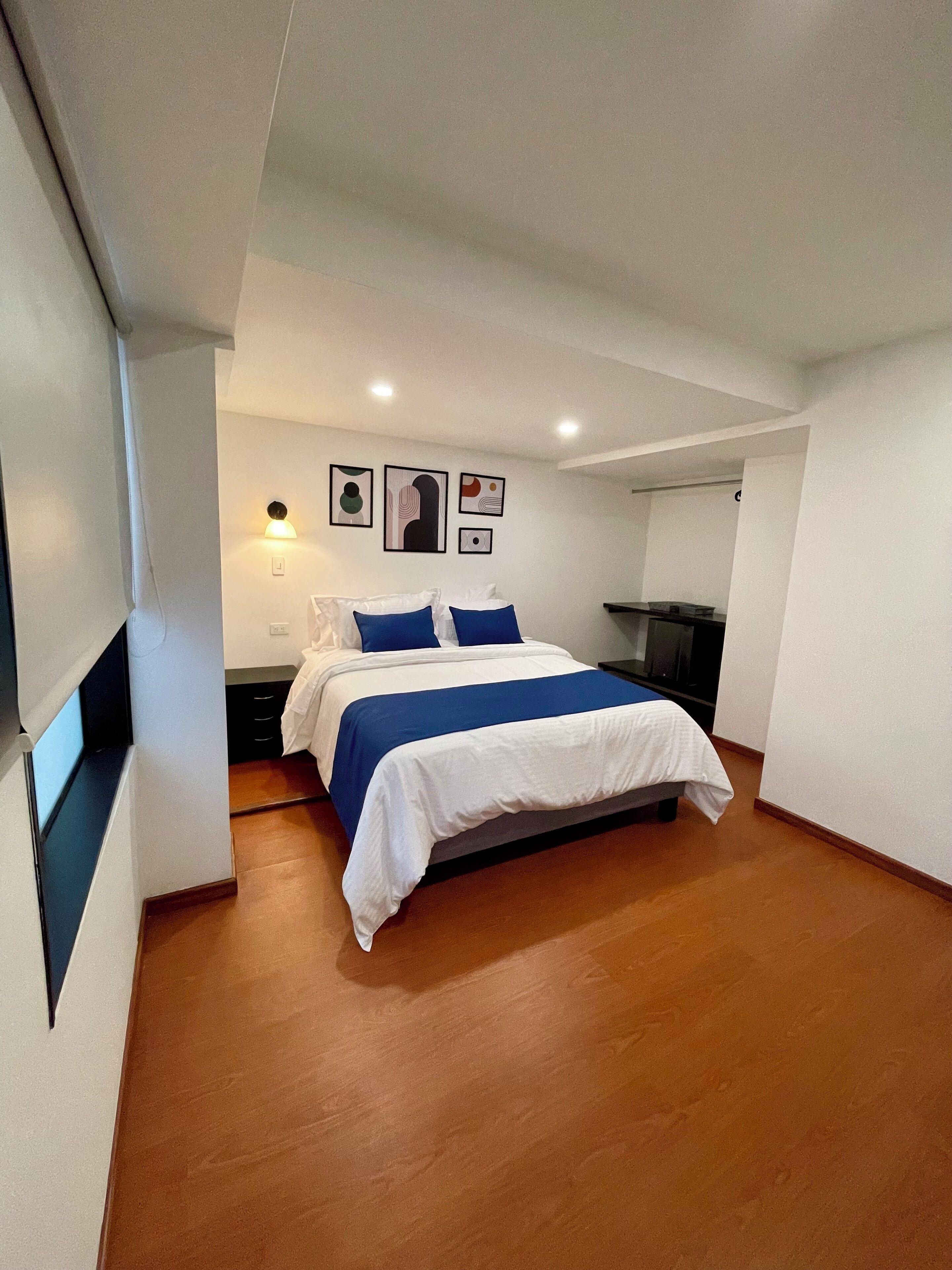 A modern bedroom with a bed, a nightstand, and a wall-mounted light. There are three framed pictures on the wall.