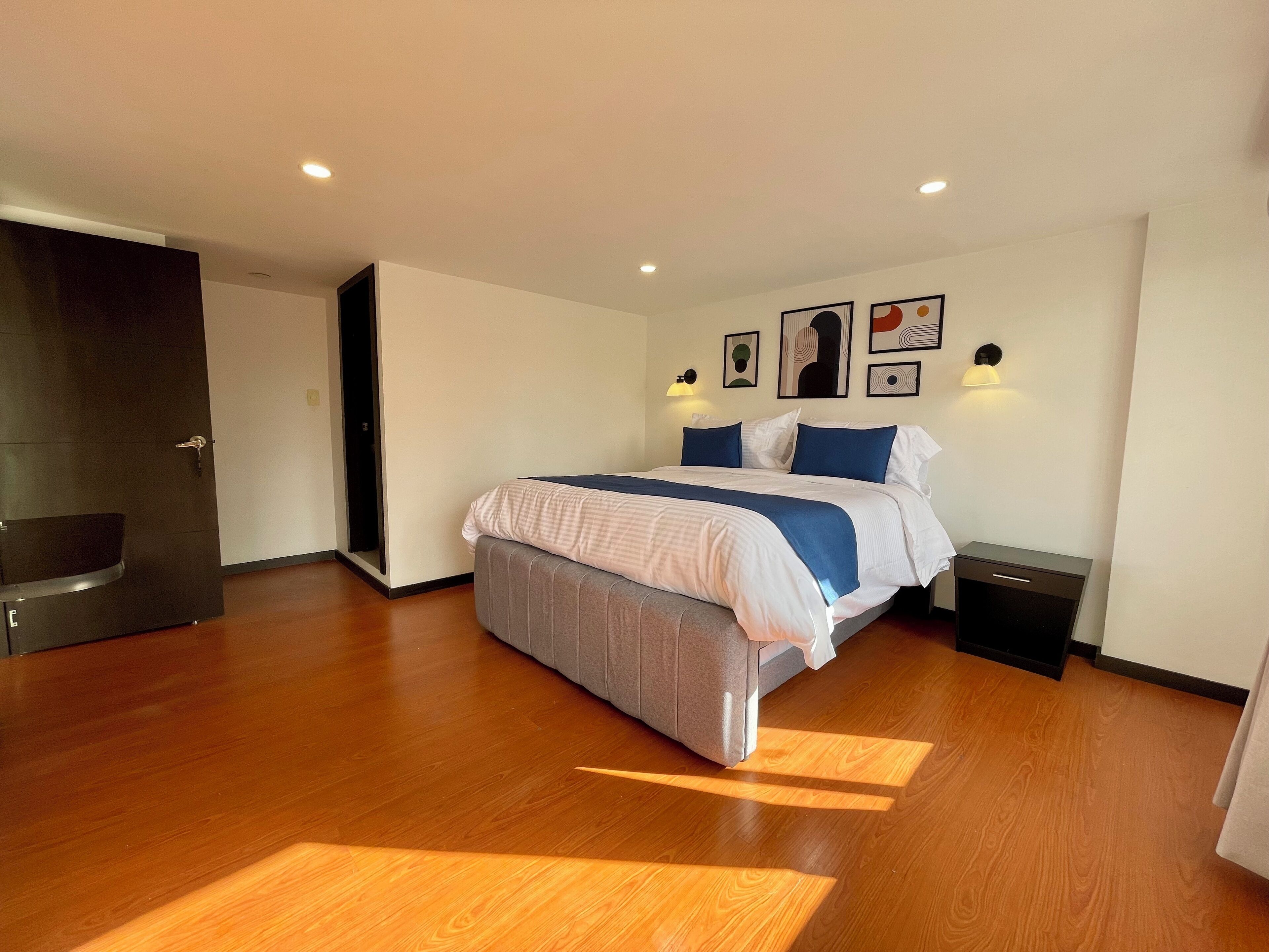 Un dormitorio moderno con una cama grande, piso de madera, una puerta oscura y luces fijadas a la pared.