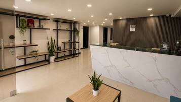 Una moderna recepción con mostrador de mármol, estanterías con plantas y un área de descanso con mesa.