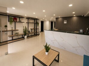 Una moderna recepción con mostrador de mármol, estanterías con plantas y un área de descanso con mesa.