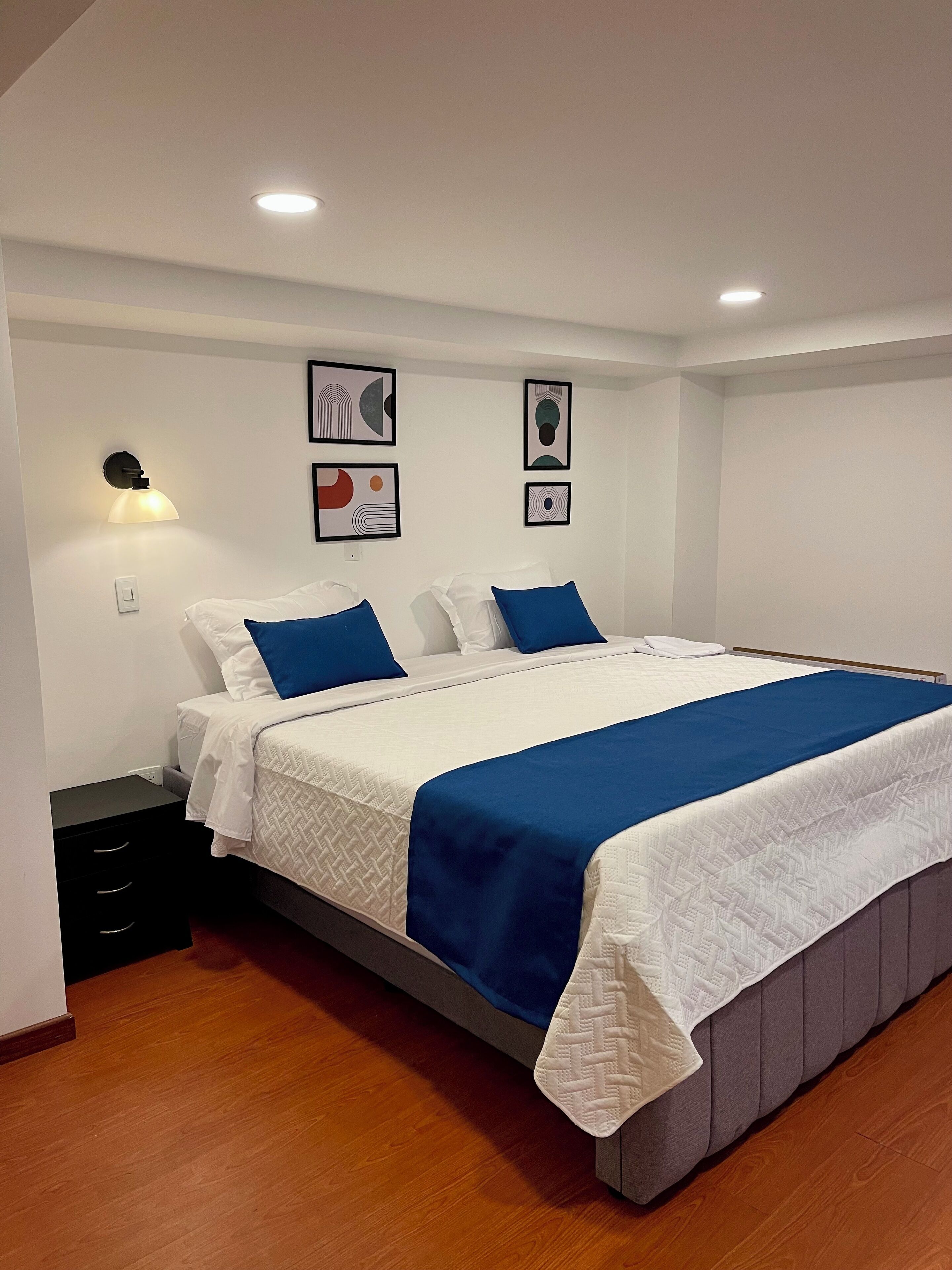 Ein ordentlich gemachtes Bett mit weißer und blauer Bettdecke, ein Nachttisch mit Lampe und Kunstwerk an der Wand.