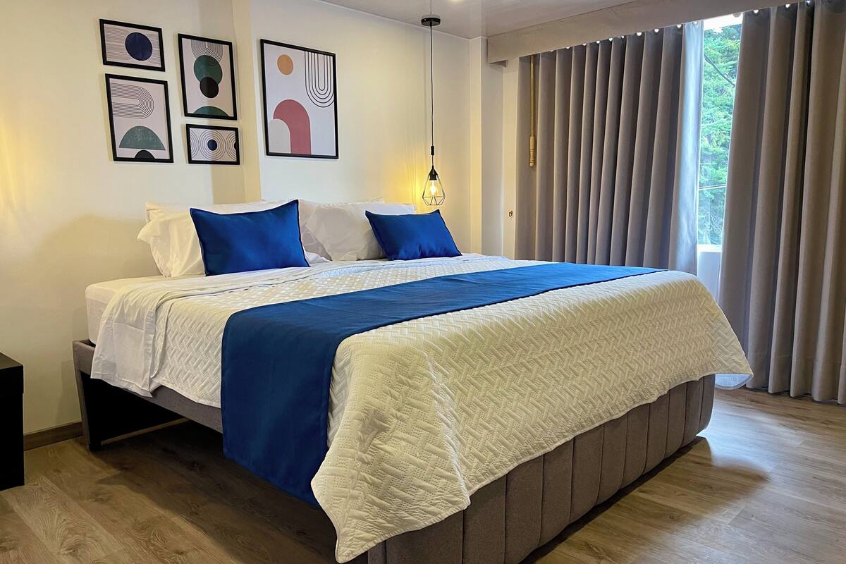 Ein ordentlich bezogenes Bett mit weißer und blauer Bettwäsche, ein hölzerner Kopfteil und ein großes Fenster mit Vorhängen.