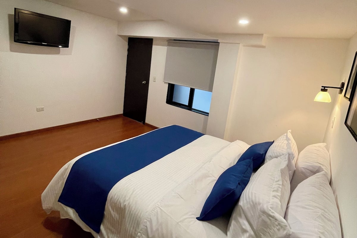 Ein Schlafzimmer mit einem Bett, einem an der Wand montierten Fernseher, einem Fenster mit Jalousie und einer Tür.