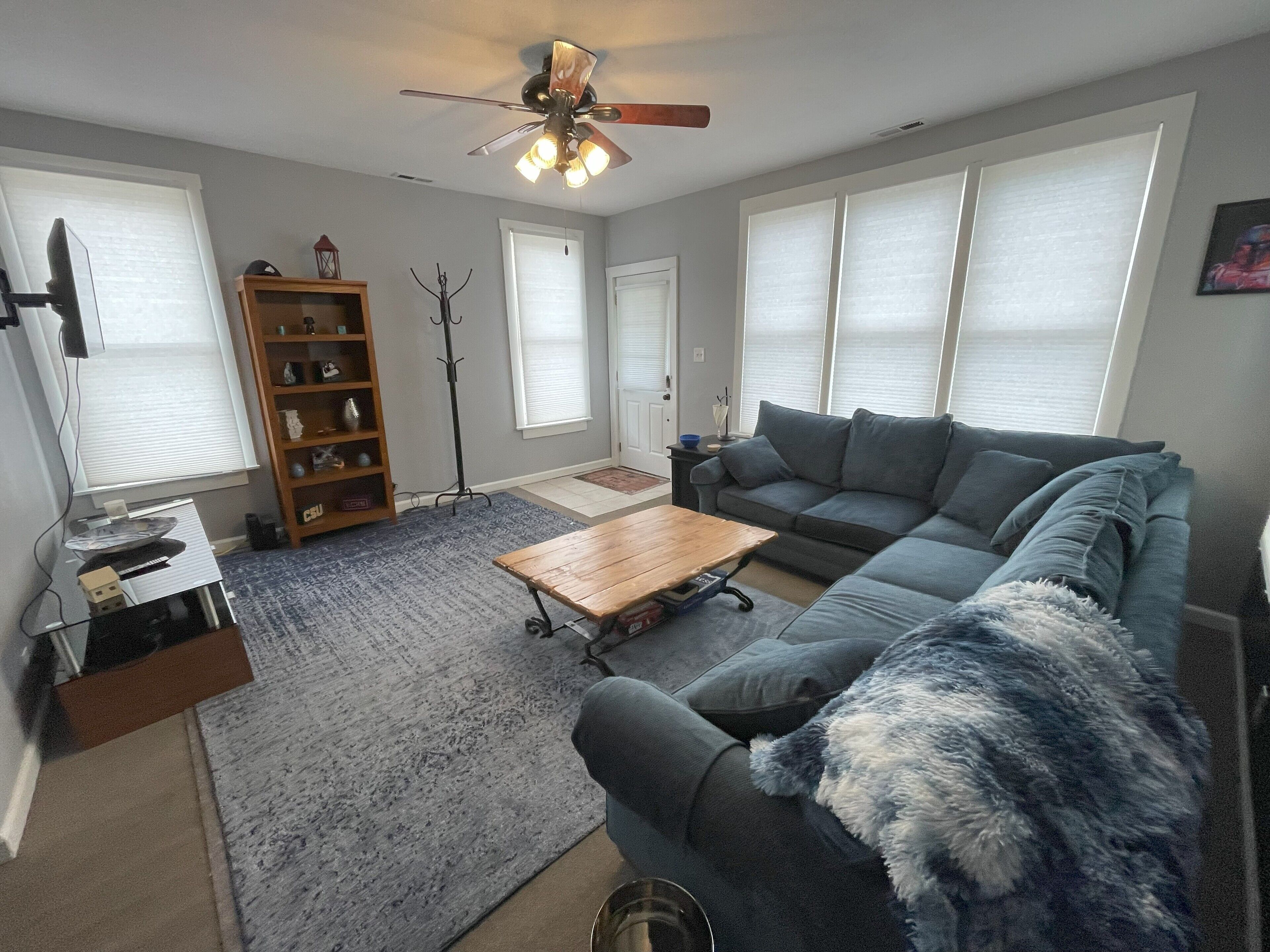 Ein Wohnzimmer mit Ecksofa, Couchtisch, Bücherregal, Stehlampe und Deckenventilator.