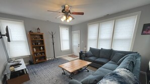 A living room with a sectional sofa, a coffee table, a bookshelf, a floor lamp, and a ceiling fan.