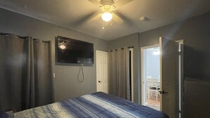 A bedroom with a bed, a ceiling fan, a TV mounted on the wall, and a door leading to a balcony.