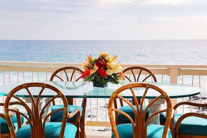 A round glass table with four wicker chairs, each with a teal cushion, set on a balcony overlooking the sea. - Private Oceanfront Home, Sleeps 10, fire pit, minutes to Kailua, backyard beach (Kailua-Kona)