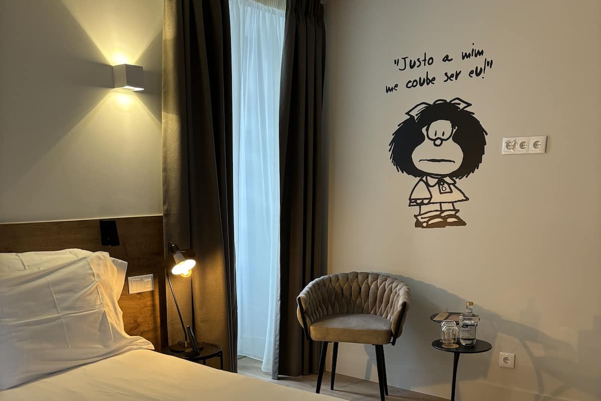 Un lit bien fait, avec du linge de lit blanc, une lampe fixée au mur, une chaise et une petite table sur laquelle se trouvent une bouteille et des verres.