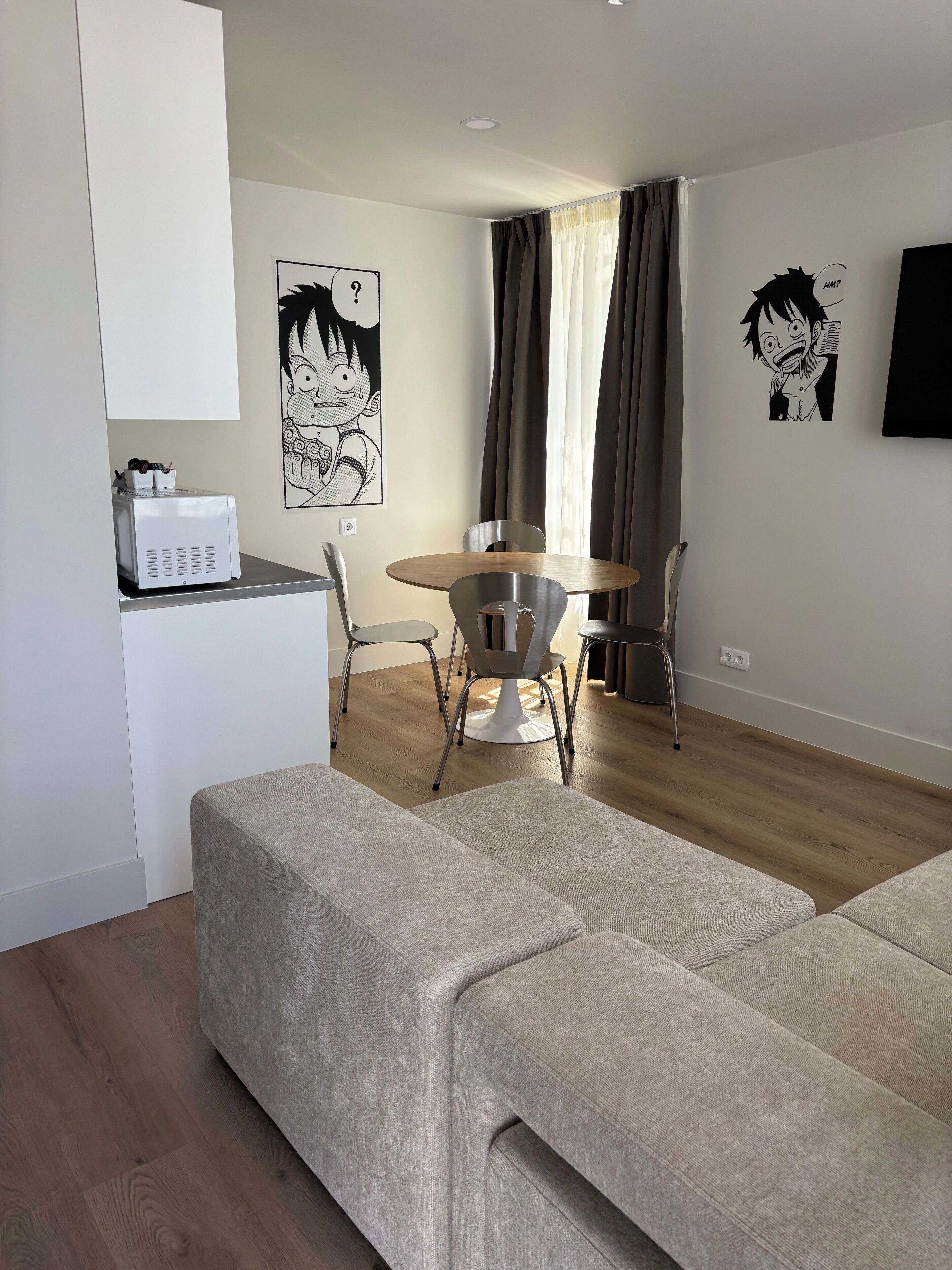 A modern living room with a sofa, a dining table with chairs, a kitchen area with a coffee machine, and two anime-themed wall decorations.