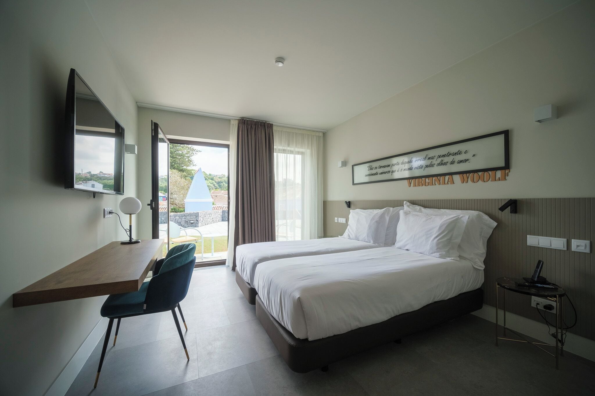 A hotel room with a large bed, a desk with a chair, a TV mounted on the wall, and a window with curtains.