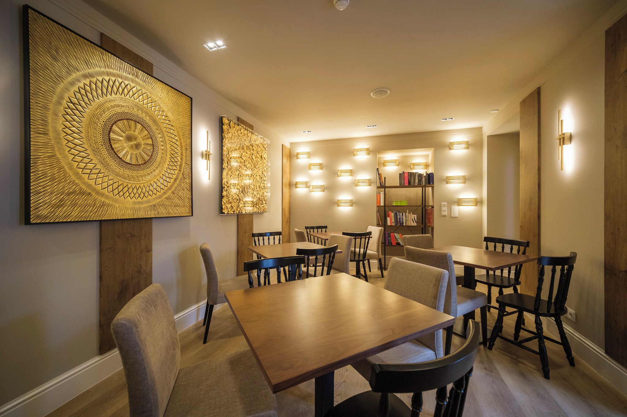 A dining area with wooden tables and chairs, a large decorative wall piece, and wall-mounted lighting.