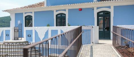 A blue building with a tiled roof, a staircase leading to a door, and a fenced courtyard.