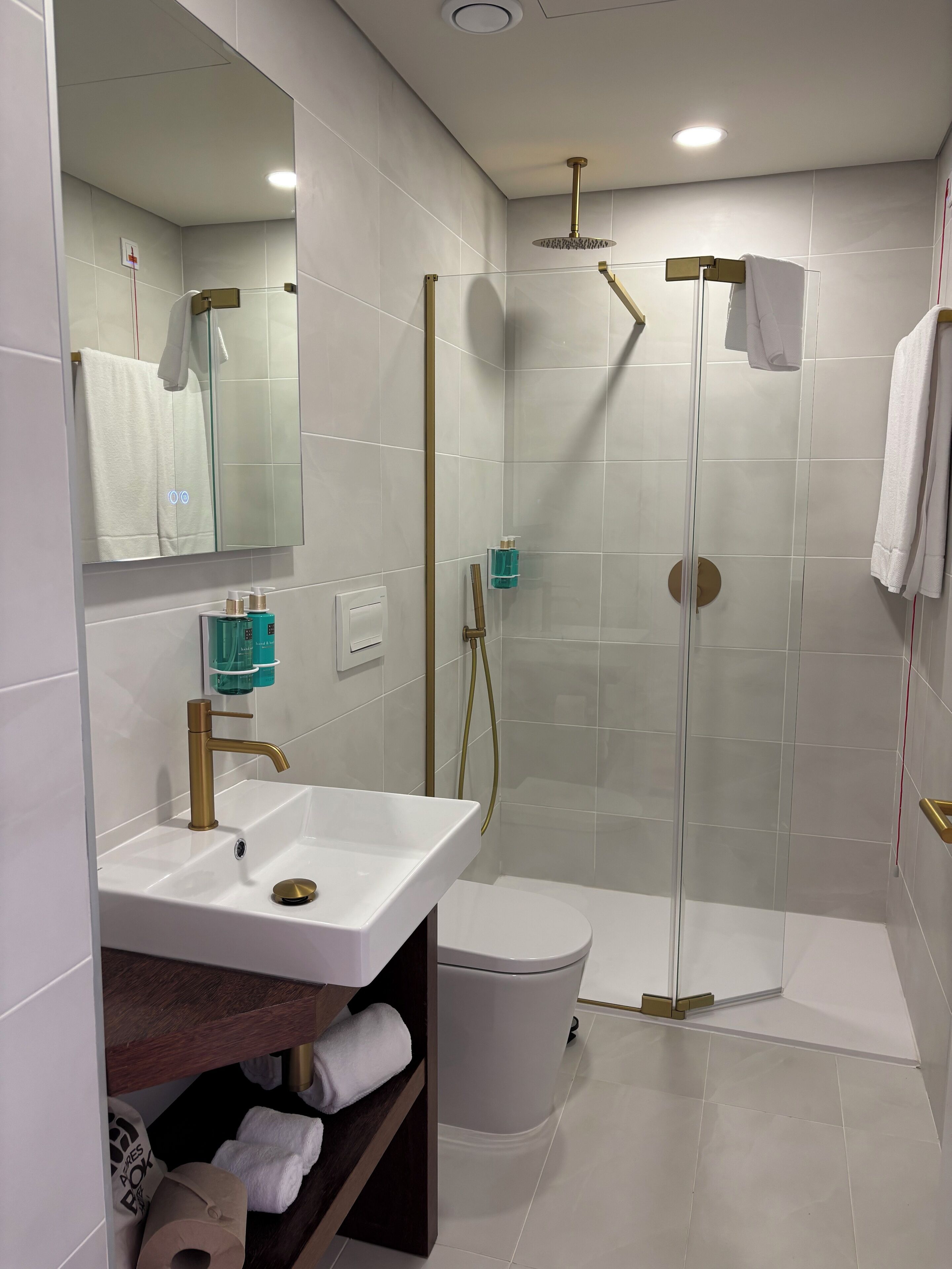 A modern bathroom with a white sink, gold fixtures, a glass-enclosed shower, and a toilet.