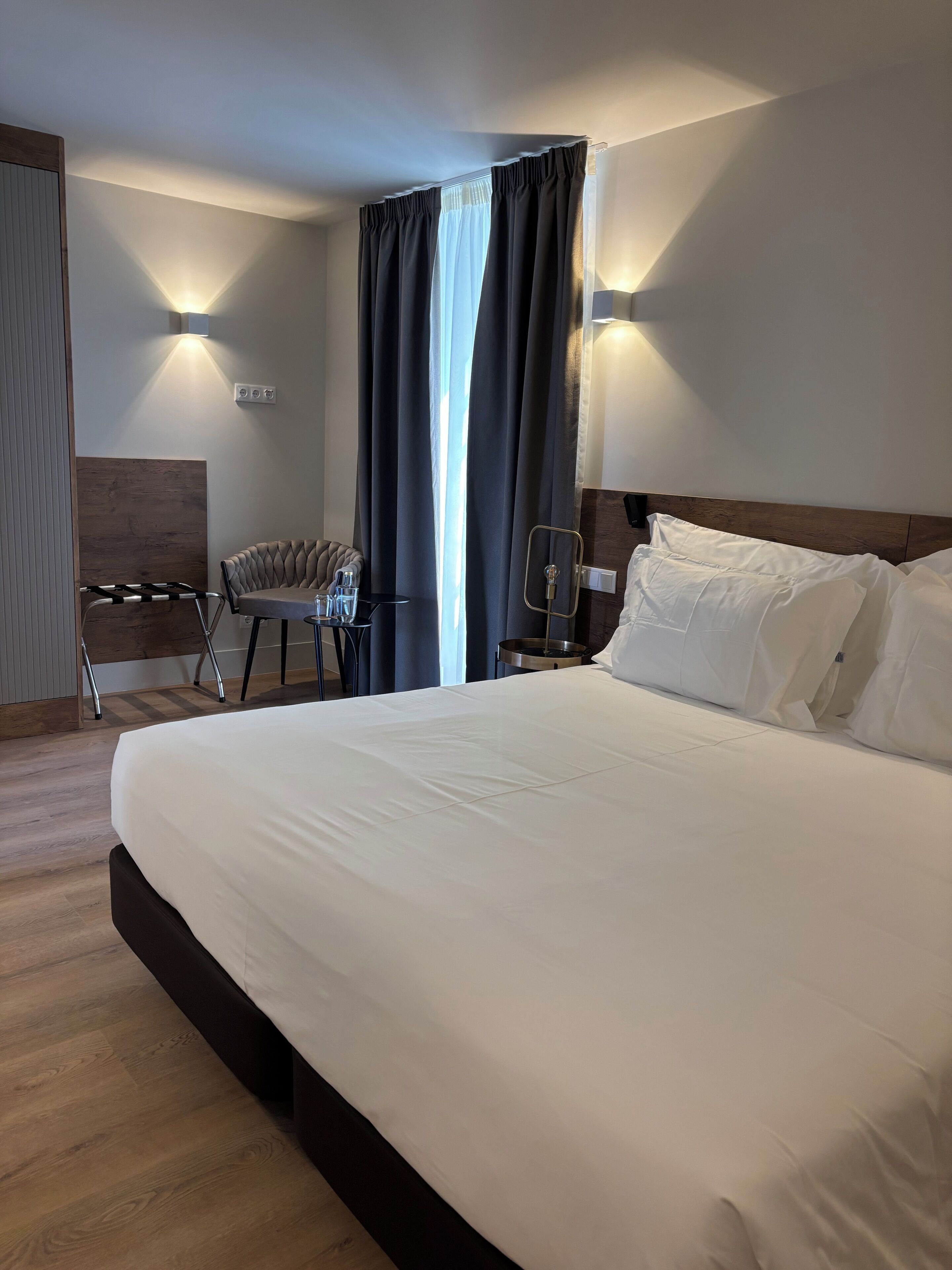 A hotel room with a large bed, a desk with a chair, a lamp, and a window with curtains.