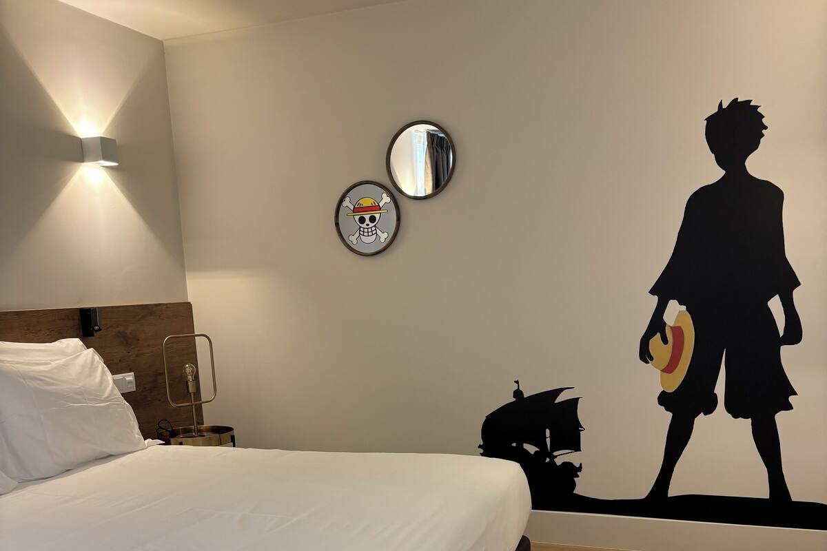 Un lit bien fait, avec du linge de lit blanc, une table de chevet avec une lampe, et un mur orné d’un miroir rond et d’une œuvre d’art représentant une silhouette.