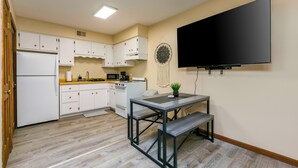 Une cuisine compacte avec des meubles blancs, un four encastré et un micro-ondes. Un téléviseur à écran plat est fixé au mur, et une petite table avec un banc se trouve au centre de la pièce.