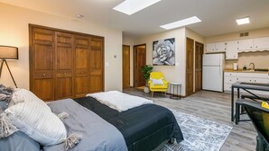 Une chambre comprenant un lit, un bureau, une chaise, une petite table et un coin cuisine avec des placards et un réfrigérateur.