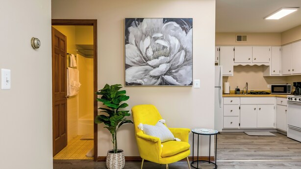 Une cuisine moderne avec des meubles blancs, un four à micro-ondes et une cuisinière. On y trouve un fauteuil jaune avec un coussin blanc, placé à côté d’un grand tableau représentant des fleurs aux tons gris et blancs, accroché au mur.