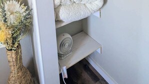 Eine Ecke eines Zimmers mit weißer Wand, Holzboden, einem weißen Regal mit weißer Decke, einem weißen Ventilator und einer Korbvase mit getrockneten Blumen.