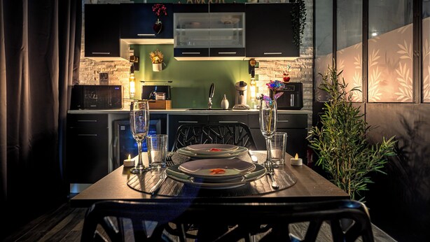 A modern kitchen with a brick wall, a green countertop, and a dining area with a table set for two. - Secret Love Room Perpignan Full Équipé et Parking Privée (Perpignan)