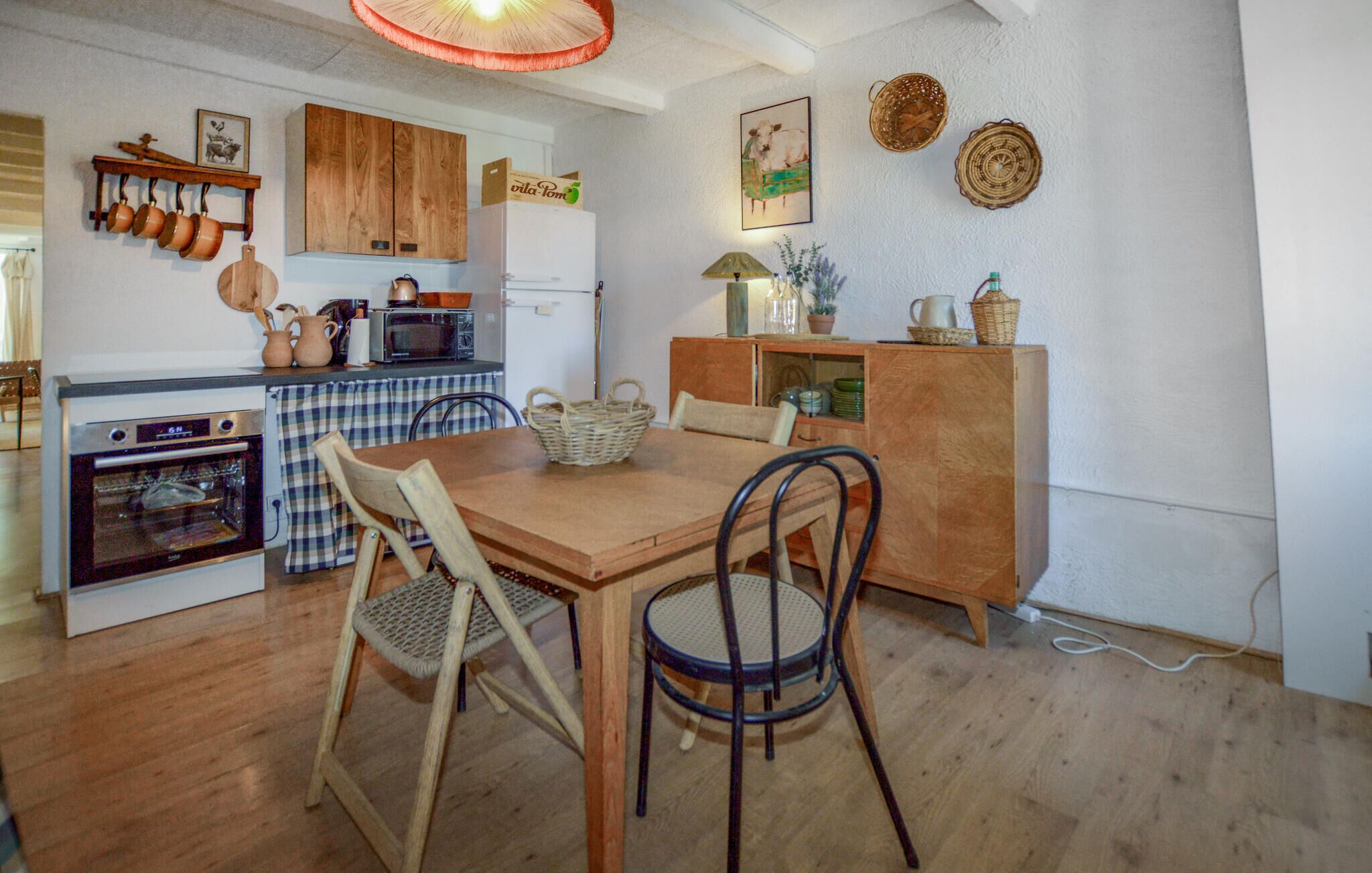 Une cuisine compacte avec des meubles en bois, un four à micro-ondes et une cuisinière. On y trouve une table à manger avec deux chaises et un petit meuble contenant des objets décoratifs.