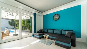 Een moderne woonkamer met een hoekbank, een klein rond tafeltje en een groot raam met uitzicht op een zwembad en groen.