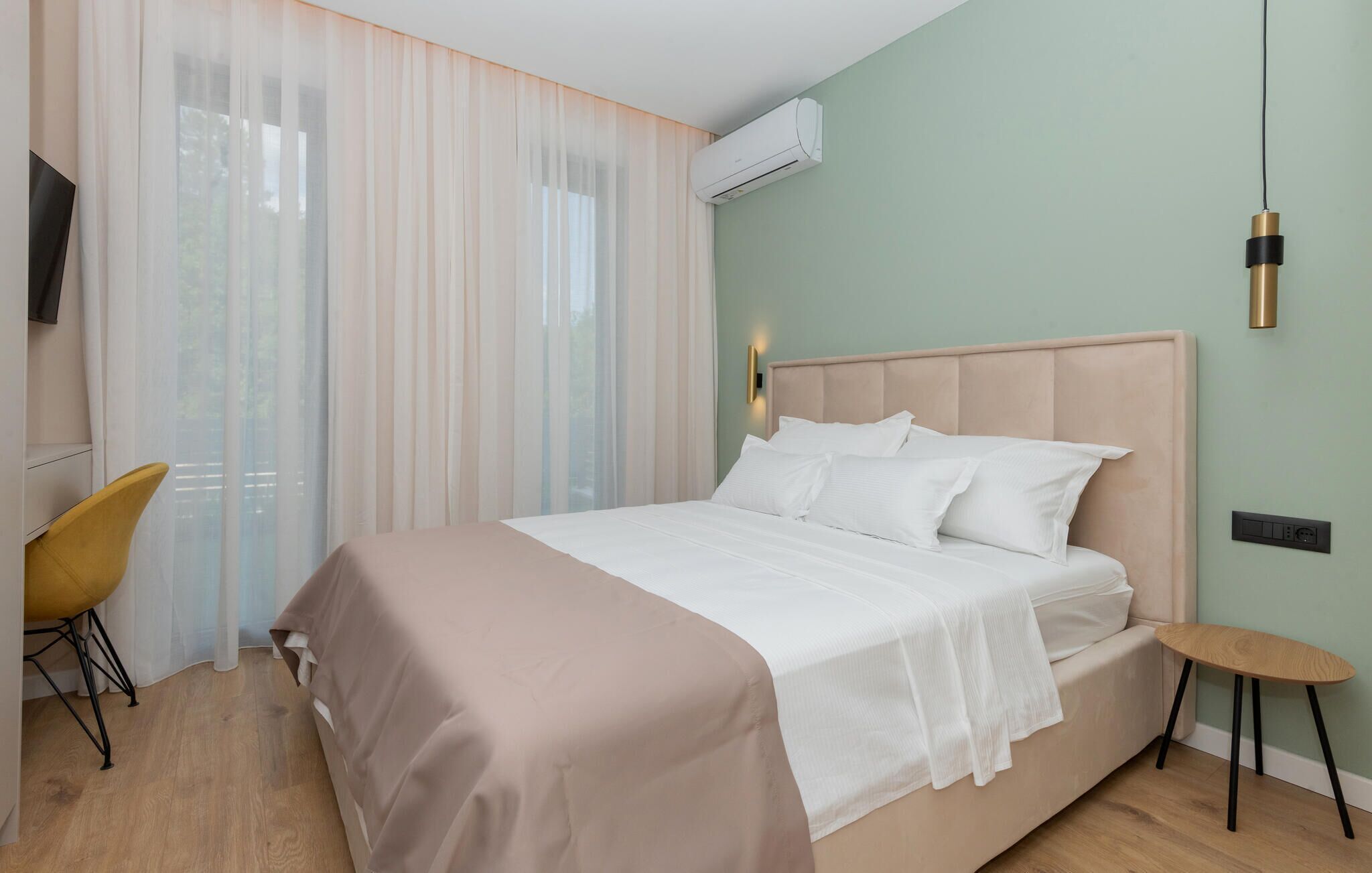 Ett hotellrum med en säng, en stol, ett litet bord, en luftkonditioneringsanläggning och en väggmonterad lampa.