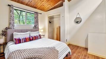 A bedroom with a wooden ceiling, a large bed with striped bedding, a window with curtains, a bedside table with a lamp, and a mirror on the wall.