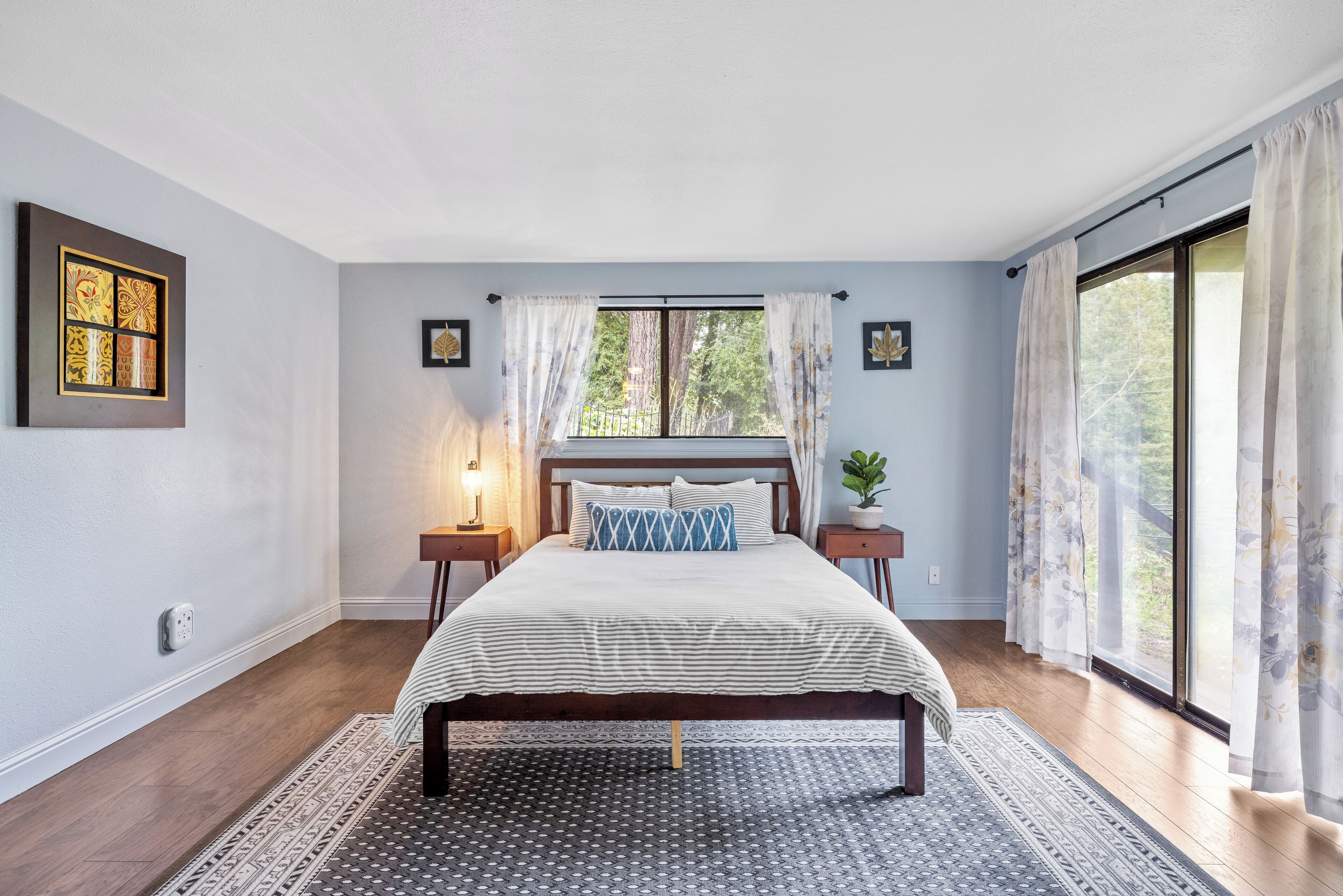 A bedroom with a large bed, a window with curtains, a bedside table with a lamp, and a wall-mounted artwork.