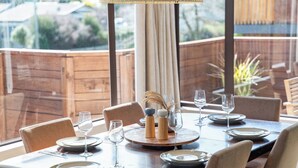 Mesa de comedor dispuesta con platos, vasos y cubiertos, con un centro de mesa decorativo y vistas al exterior a través de amplias ventanas.