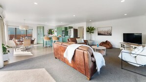Una sala moderna con un sofá de cuero marrón, un comedor con mesa y sillas, una cocina con armarios verdes y un ventanal con vistas.